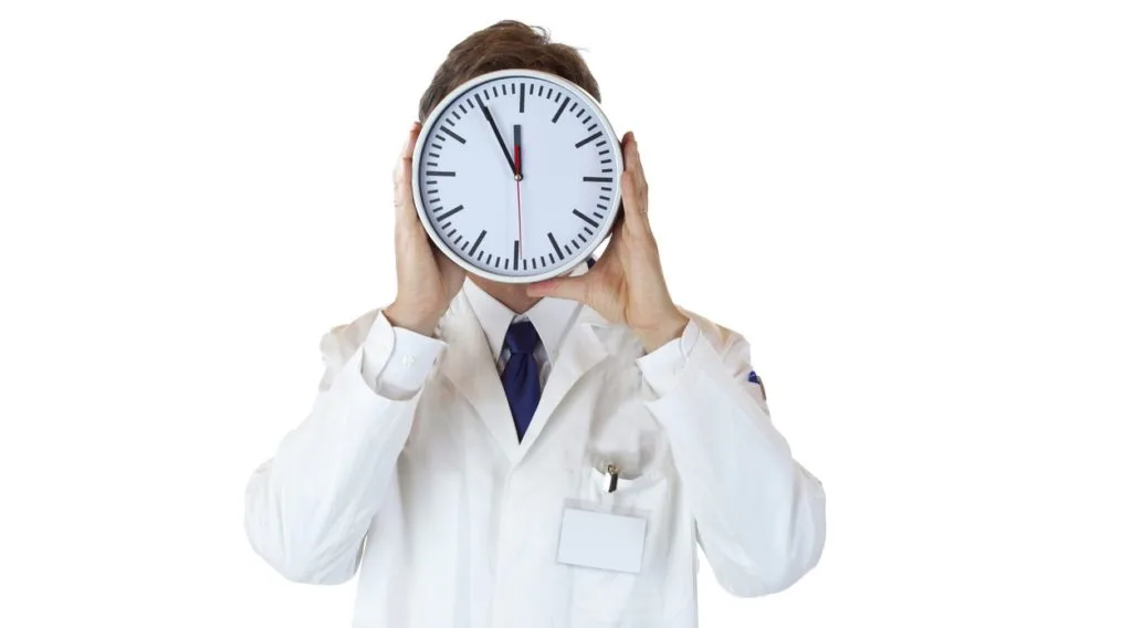 A doctor looking stressed in front of a clock in an office.