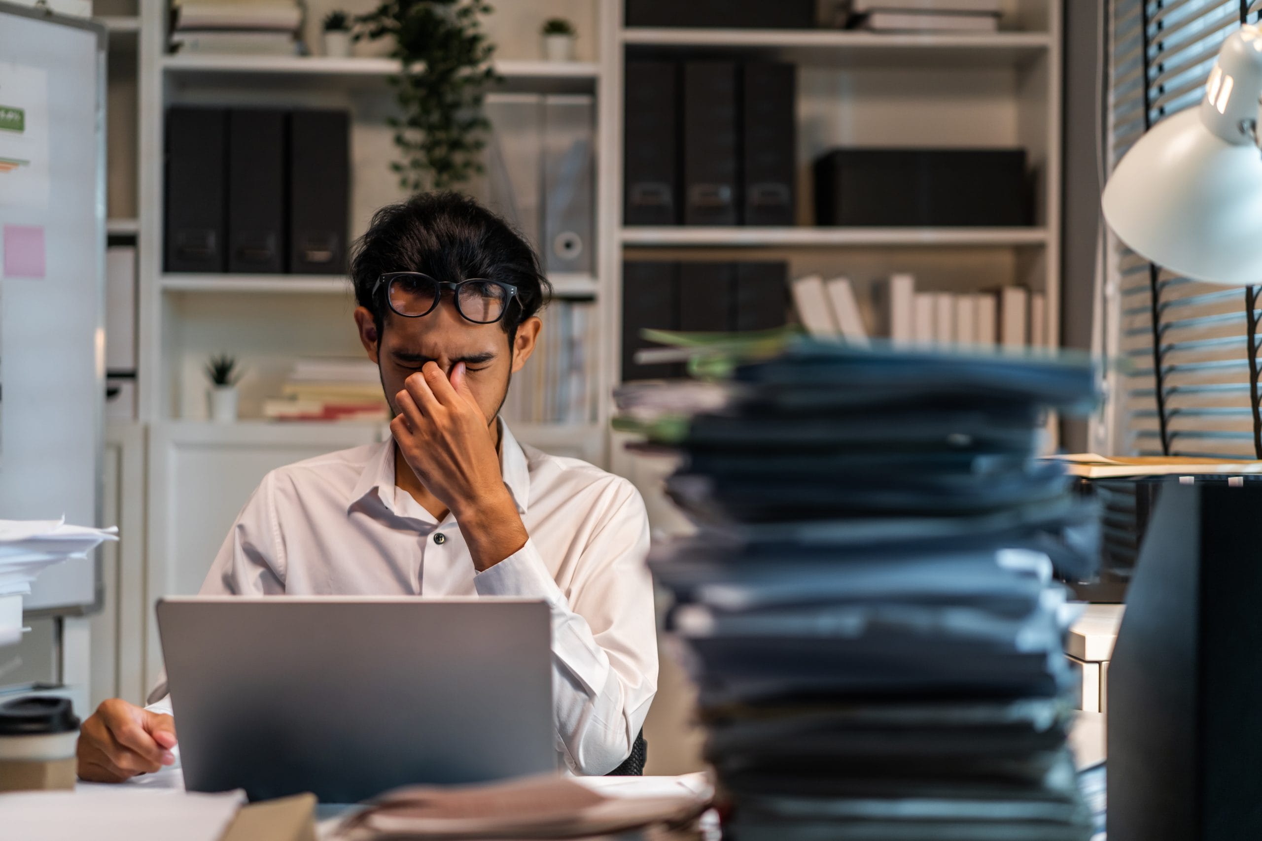 A man behind a desk with piles of paperwork, looking overwhelmed