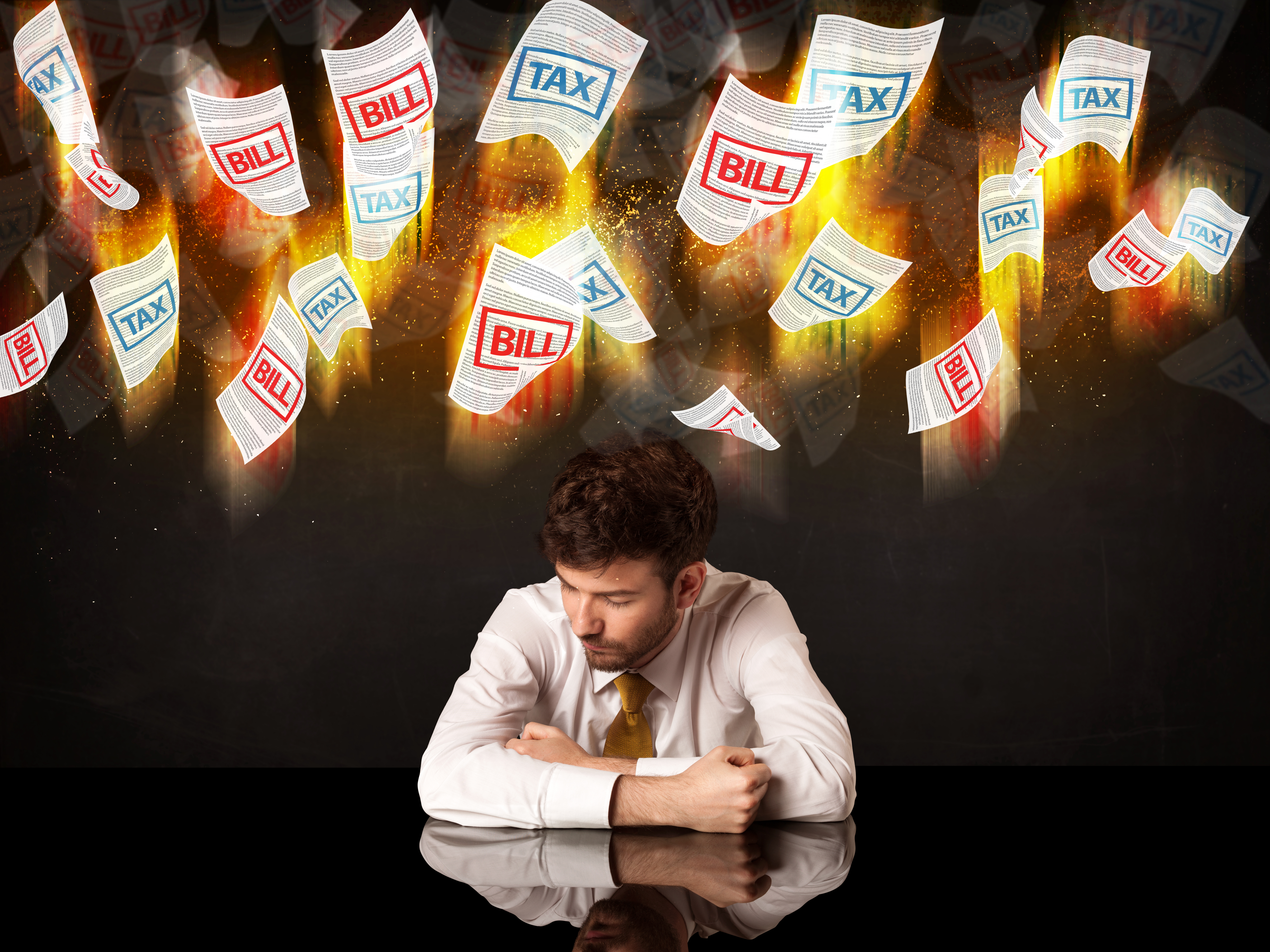 a male sitting with his head close to the tables and pieces of flaming paper with the words bills and tax flying above his head